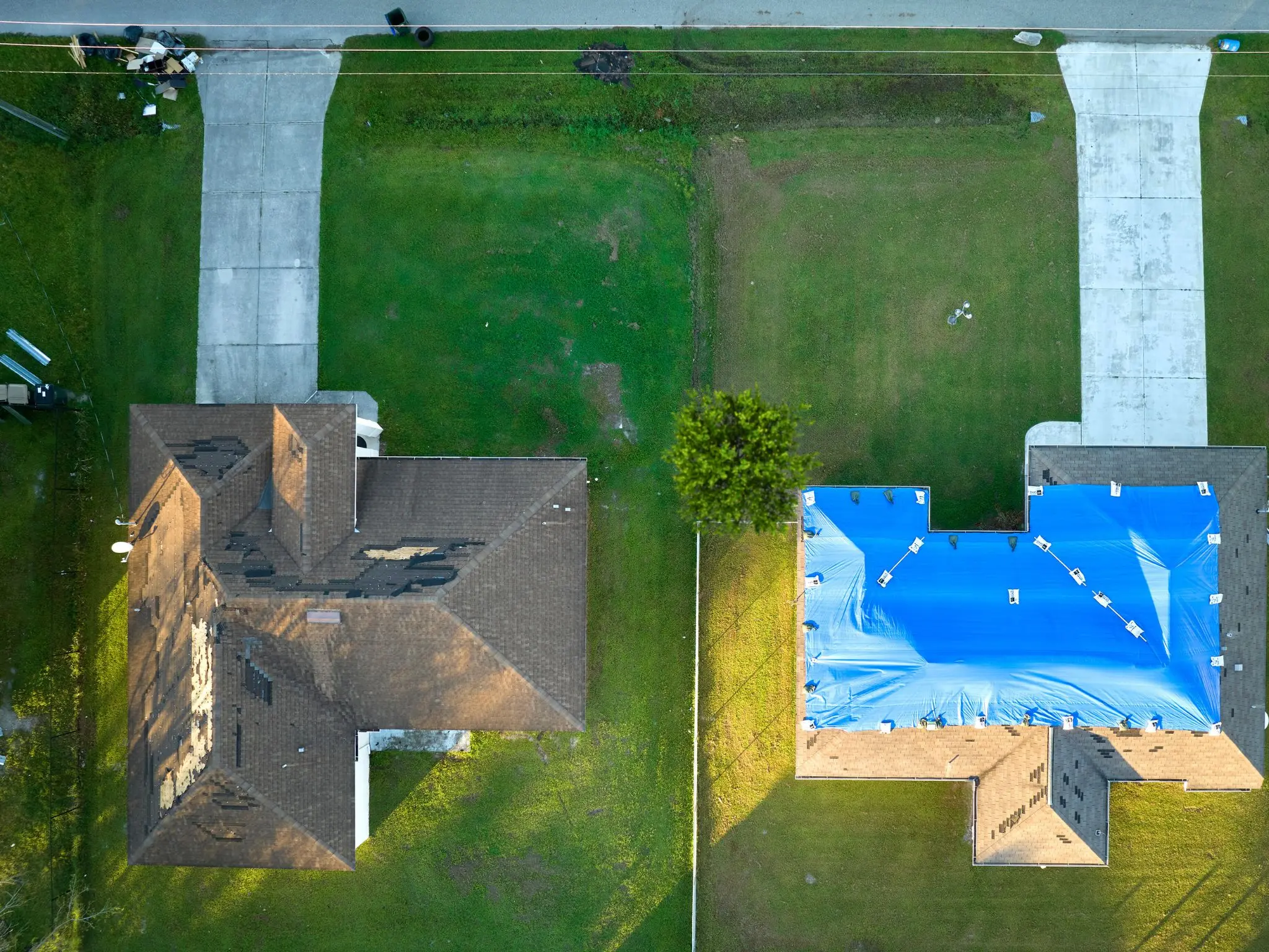 drone shot of storm damaged home roofs. one visibly missing shingles and one covered with blue tarp to protect from further damage.